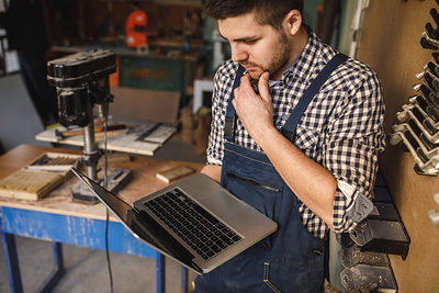 Young carpenter working on a laptop in his workshop. Schlagwort(e): workshop, carpentry, occupation, carpenter, laptop, professional, craftsman, handyman, industry, craft, job, timber, lumber, woodwork, handmade, tools, old, business, manual, worker, plan, expertise, computer, pursuit, art, person, careers, equipment, activity, craftsmanship, talent, decoration, lifestyle, wooden, classic, handicraft, design, skill, capability, career, style, networking, working, woodworker, online, using, smiling, workman, indoors, workshop, carpentry, occupation, carpenter, laptop, professional, craftsman, handyman, industry, craft, job, timber, lumber, woodwork, handmade, tools, old, business, manual, worker, plan, expertise, computer, pursuit, art, person, careers, equipment, activity, craftsmanship, talent, decoration, lifestyle, wooden, classic, handicraft, design, skill, capability, career, style, networking, working, woodworker, online, using, smiling, workman, indoors